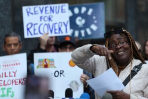 Woman in glasses points to peice of paper in front of a crowd holding poster signs, one of which says "Revenue for Recovery"