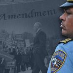 Police officer in focus with protestors in the background holding "Stand with Mark" signs. The 1st Amendment is written across the top of the image.
