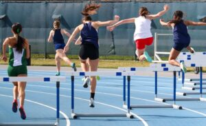 Five female athletes participating in a hurdle race on a running track.